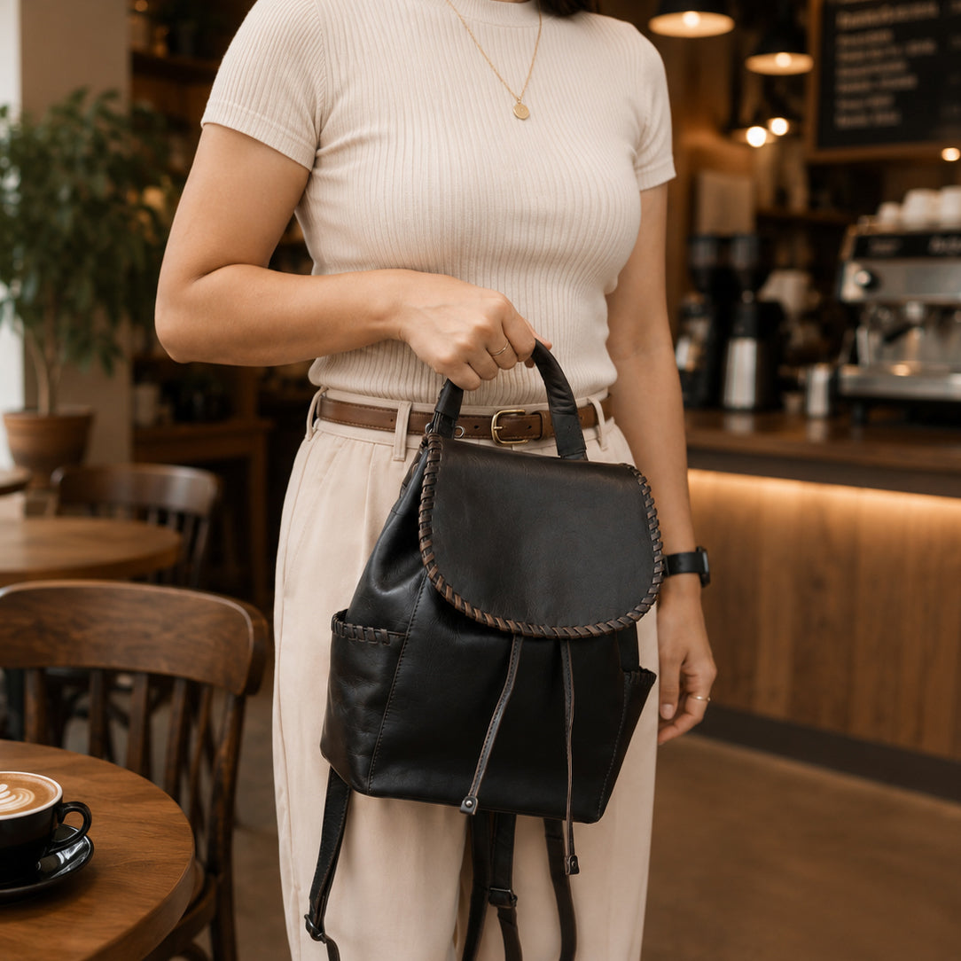 A woman in a cream ribbed short-sleeve top and beige trousers holds a black leather backpack with braided trim in a cozy, warmly lit cafe setting.