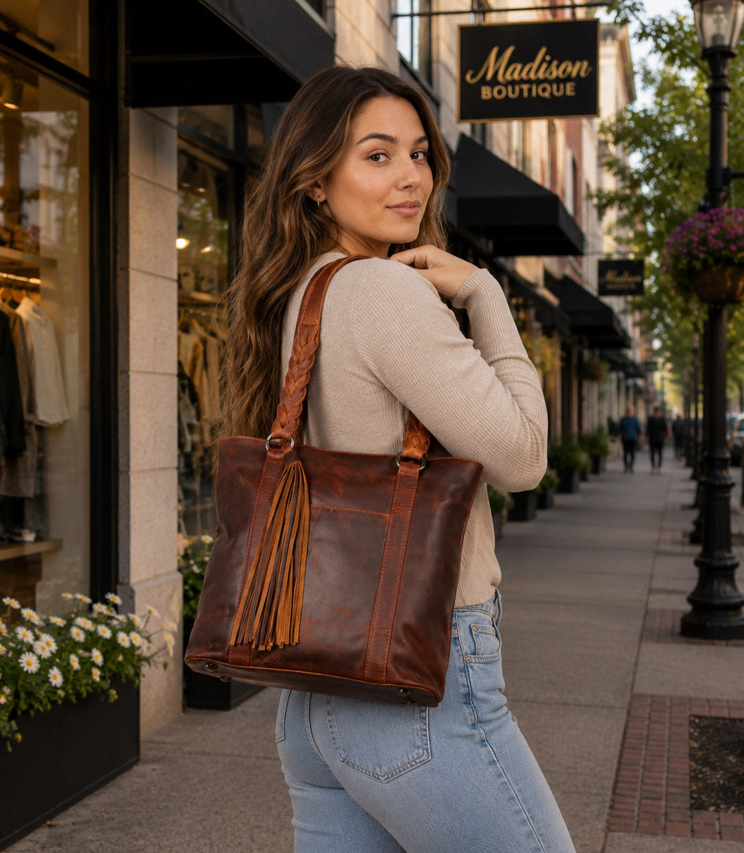A woman wearing a beige long-sleeve top and light blue jeans carries a large brown leather tote bag with tassel fringe on a city sidewalk carrying The Bella Concealed Carry Tote in Cognac