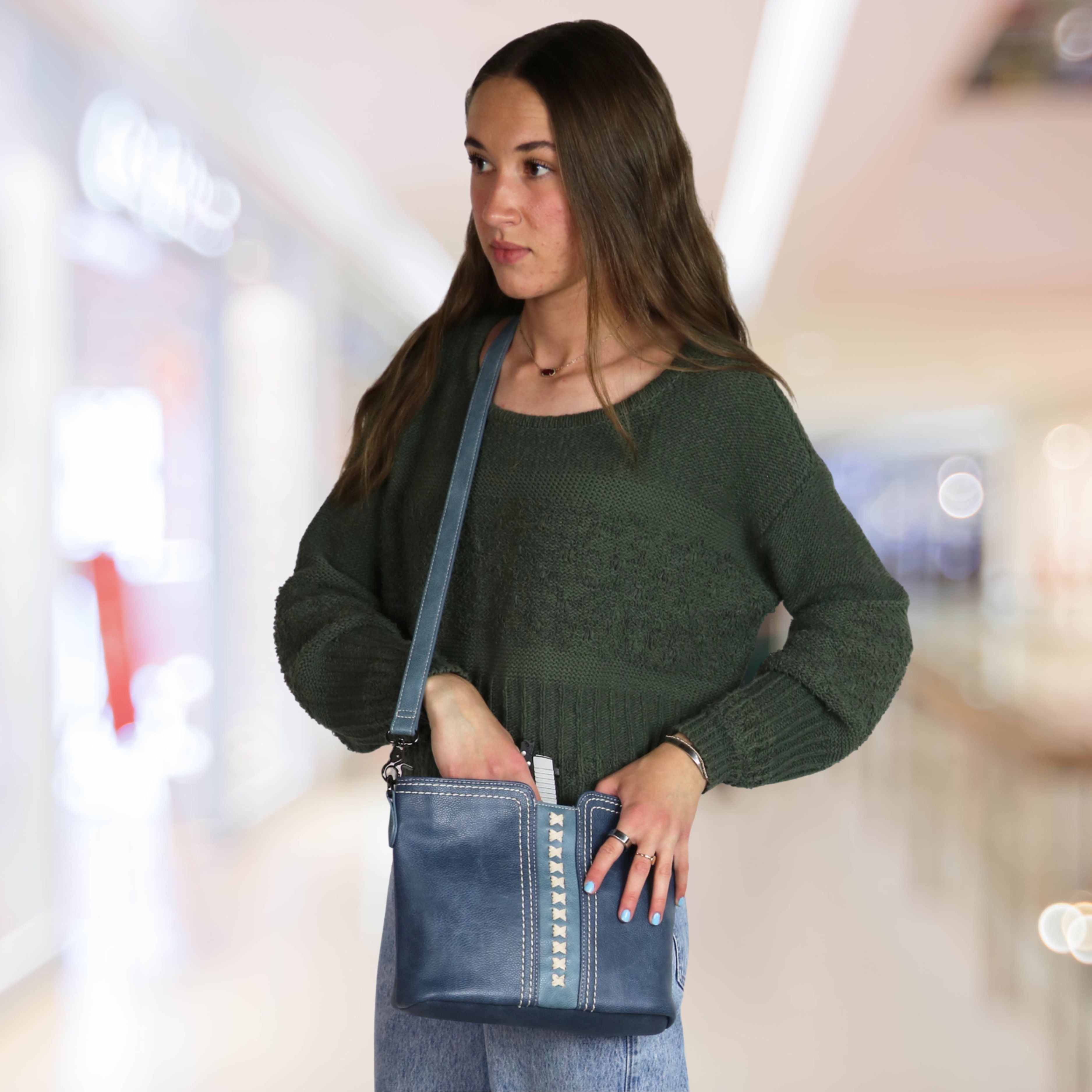A woman wearing a dark green knit sweater and jeans carries a blue leather shoulder bag with silver studs in a blurred indoor shopping mall setting.