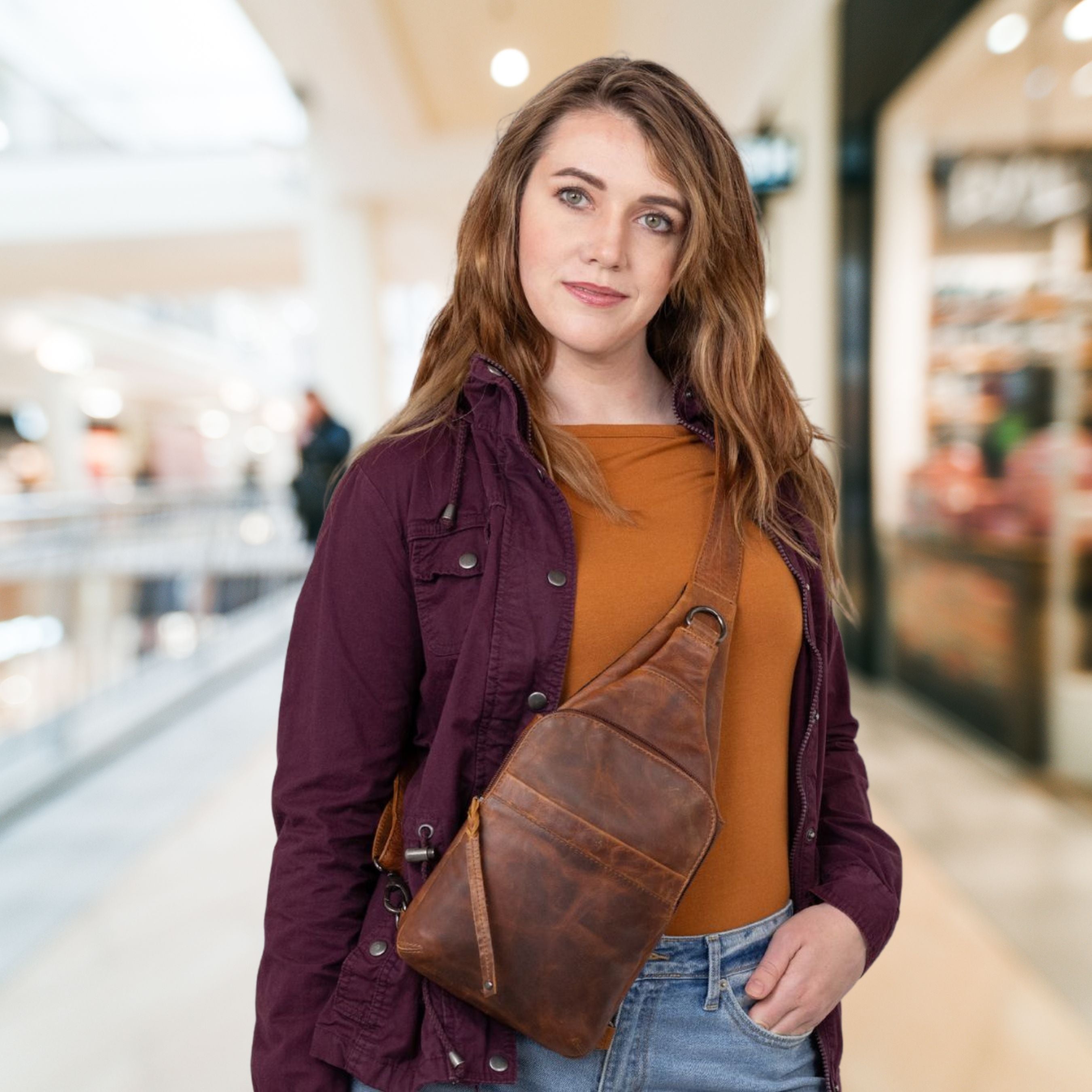 A woman wearing a brown leather Dakota sling bag across her torso, standing in a brightly lit, blurred indoor shopping mall environment.