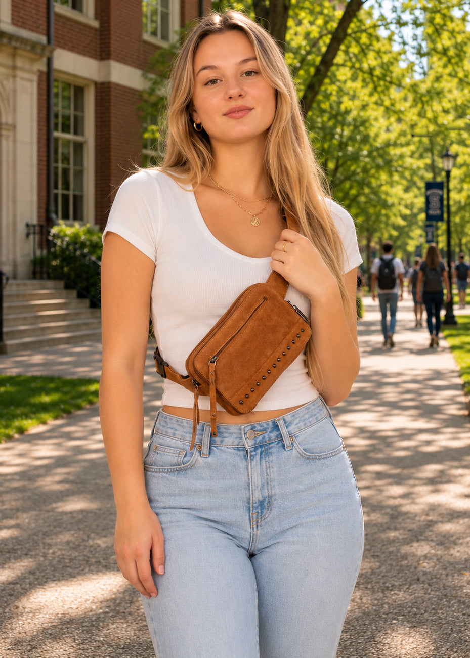 A young woman stands on a sunlit campus walkway wearing a white cropped T-shirt, light blue jeans, and the Kailey concealed carry brown suede crossbody bag with a zipper and tassel detail.