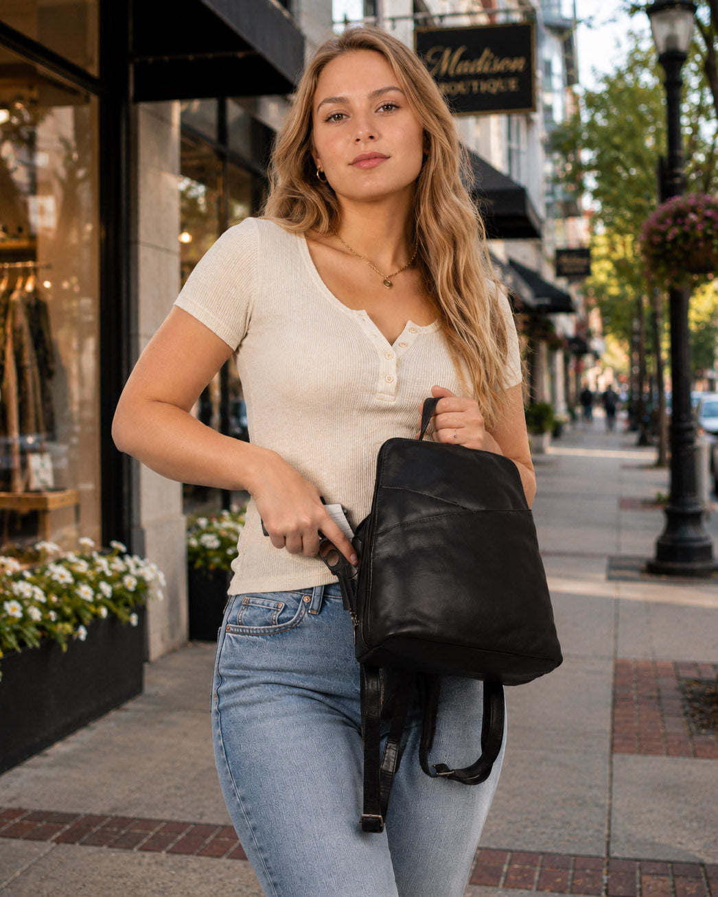 A woman walking on a city sidewalk wearing a cream short-sleeve henley top and light blue jeans, carrying a black leather backpack.