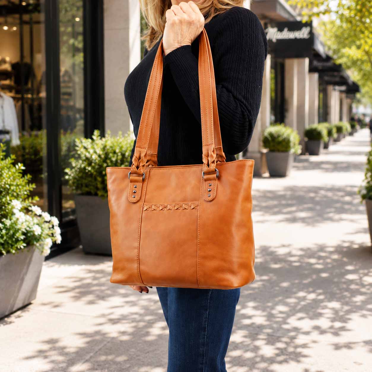 A woman carrying a large tan leather tote bag on a sunny outdoor sidewalk. The bag has long shoulder straps, a structured rectangular shape, and decorative stitching across the front pocket area.