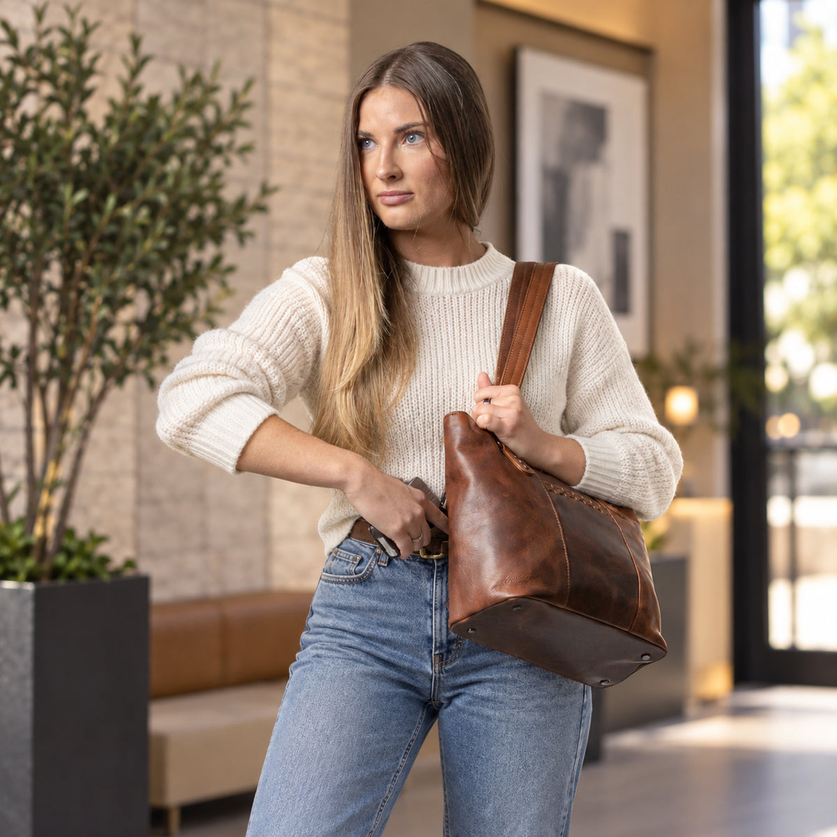 A woman wearing a cream ribbed knit sweater and blue jeans carries a large brown leather shoulder bag in a modern indoor setting. Women Lady Conceal -  YKK Locking & Universal Holster -  Designer Luxury Leather Carry Handbag -  carry Handbag for gun carry -  Unique Tote gun Handbag 
