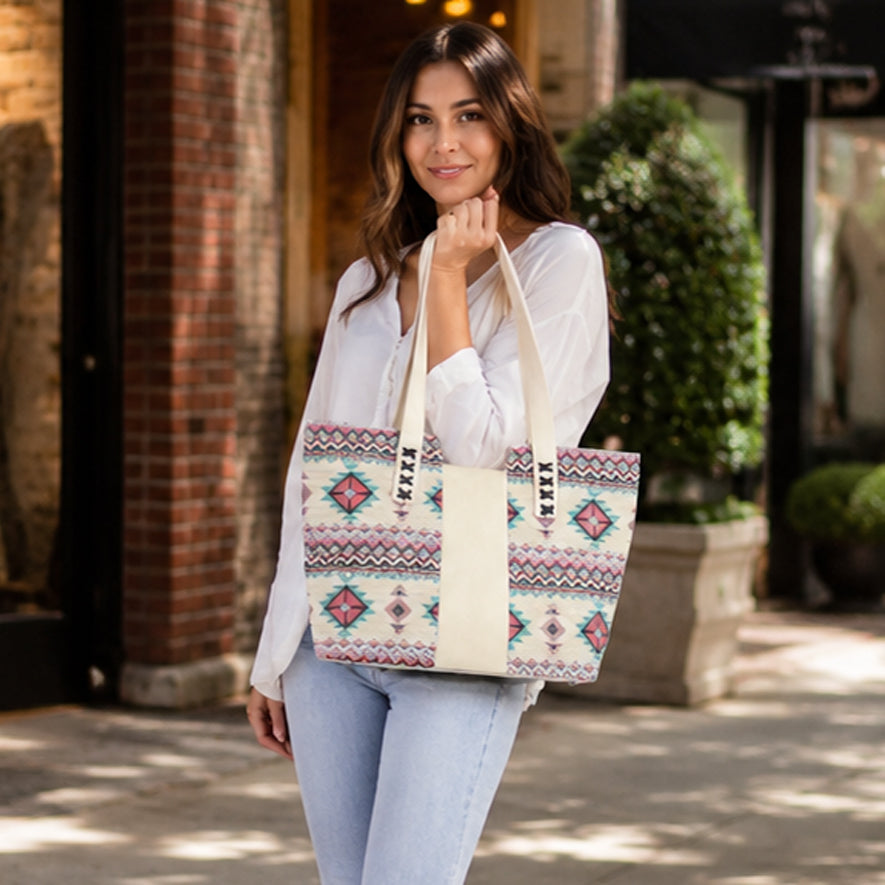 A woman wearing a white blouse and light blue jeans carries a large tote bag with a cream base and colorful geometric Southwestern-style patterns in pink, blue, and red. The brand name 'SARAH VALE' is printed vertically on the handles.